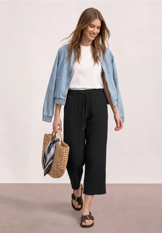 Vrouw in wit shirt, zwarte culottes, spijkerjasje, strooien tas en sandalen voor een lichte achtergrond.