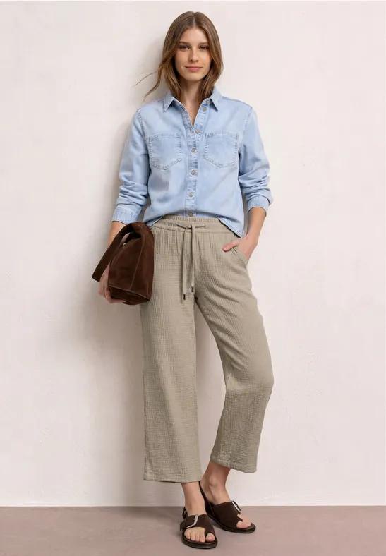 Model draagt lichtblauw overhemd, grijsbruine culotte, donkere sandalen en houdt een bruin accessoire vast.