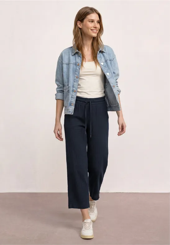 Vrouw in wijde donkerblauwe broek, wit topje en spijkerjasje, witte sneakers tegen een neutrale achtergrond.