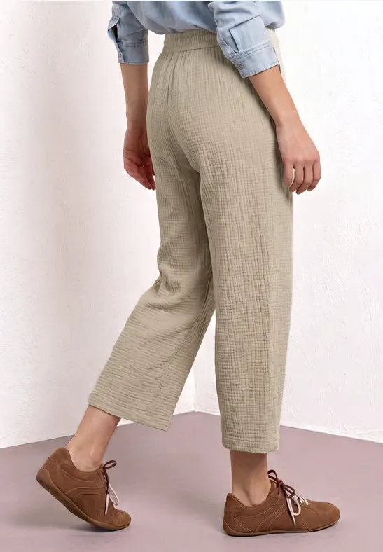 Lichtbeige broek met hoge taille en wijde pijpen tot net boven de enkel, gecombineerd met bruine veterschoenen.