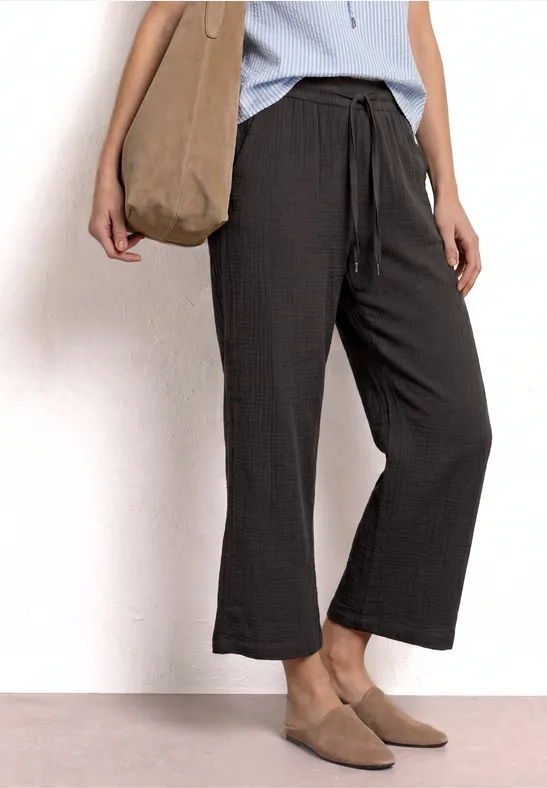 Vrouw in lichtblauw topje, zwarte wijdvallende 7/8-broek, beige mules en een stoffen tas.