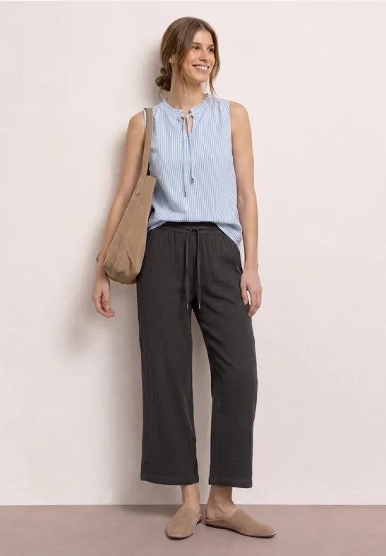 Een vrouw in lichtblauwe mouwloze blouse met strik, wijde donkergrijze broek, beige slippers en een tas.