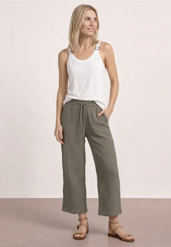Vrouw in wit shirt, losse linnen broek en sandalen, ontspannen pose tegen neutrale achtergrond.