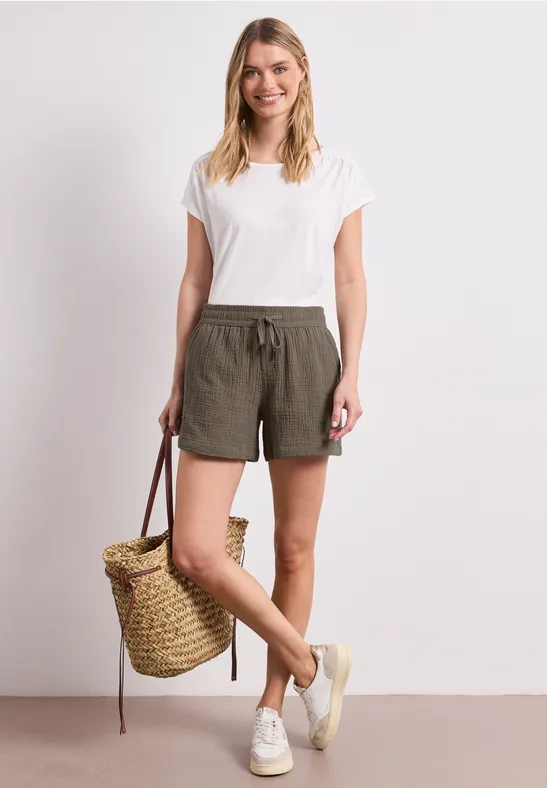 Frau in weißem T-Shirt und olivfarbenen Shorts, hält einen Korb und trägt weiße Sneaker.