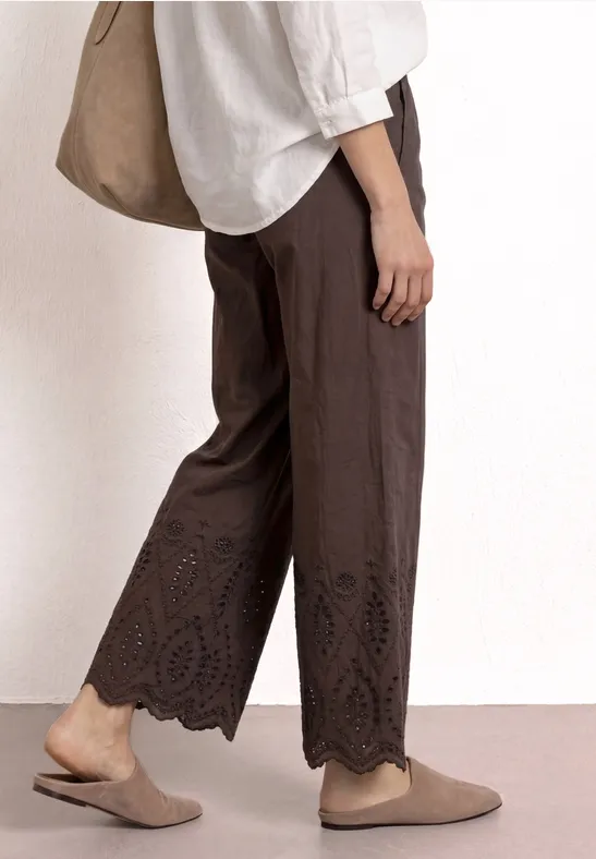 Seitenansicht einer Person mit weiten, braunen Hose mit Spitzensaum, weißem Oberteil, beigen Slippern und Tasche.