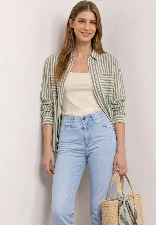 Eine Frau in hellblauer Jeans, weißem Top, gestreiftem Hemd und Korbtasche vor hellem Hintergrund.