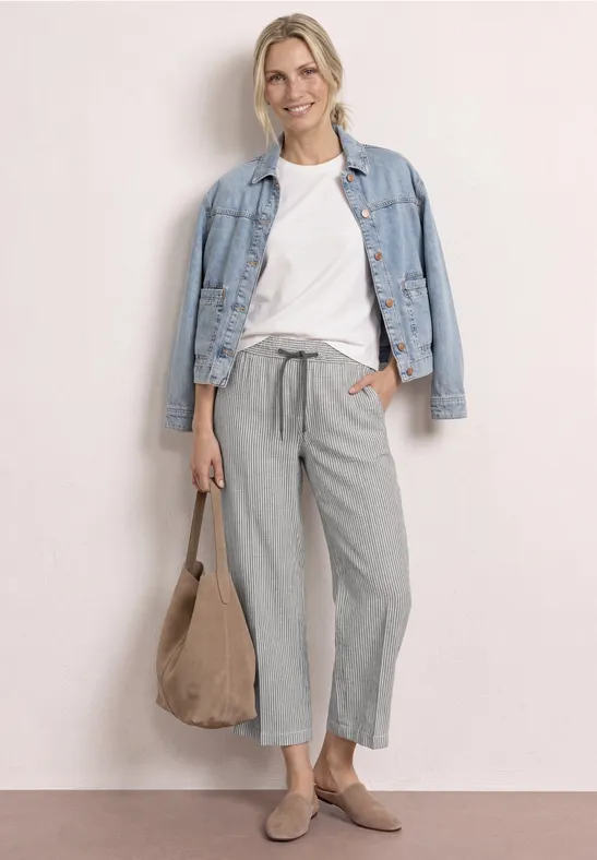 Vrouw draagt wit T-shirt, lichtblauwe spijkerjas, grijze wijde broek, beige suède tas en instappers.