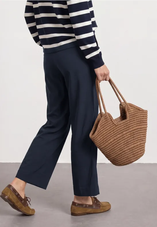 Persoon met gestreepte trui, wijde donkerblauwe broek, gevlochten handtas en bruine plateau loafers.