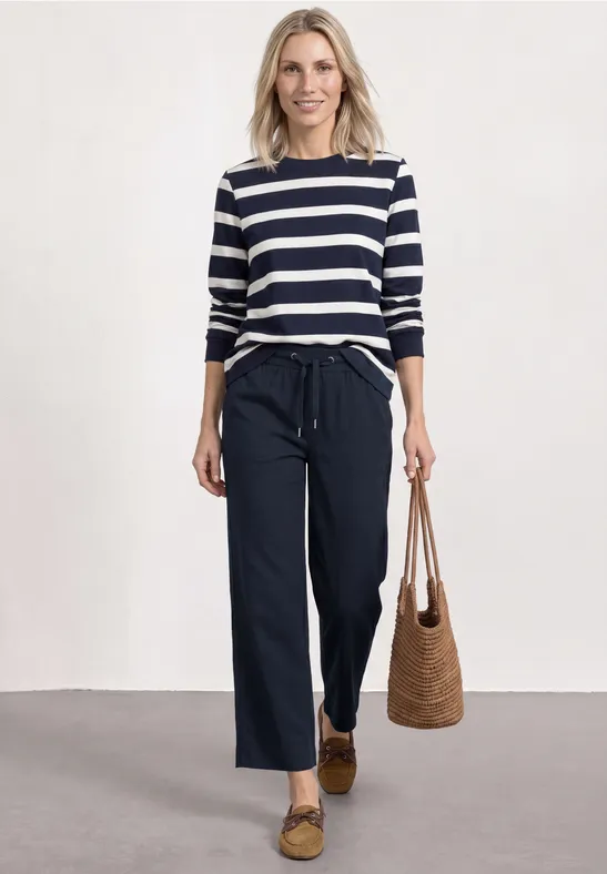 Vrouw met blauw-wit gestreept shirt, wijde donkere broek, rieten tas en loafers.