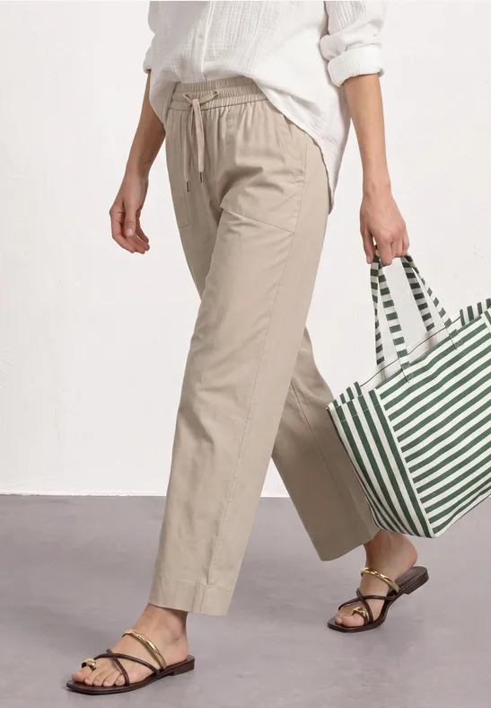 Benen van een persoon met beige broek, sandalen, wit overhemd en een groen-wit gestreepte boodschappentas.