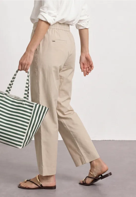 Persoon in beige broek en wit overhemd, met gestreepte groen-witte tas en platte sandalen.