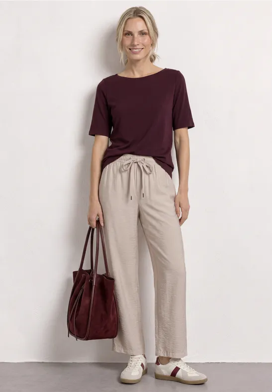 Vrouw voor witte achtergrond, met een bordeaux T-shirt, beige broek, witte sneakers en een bordeaux tas.