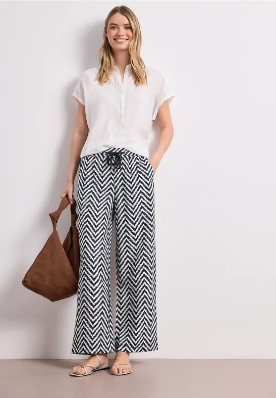 Vrouw in witte blouse, zwart-witte zigzagbroek, bruine tas en sandalen.