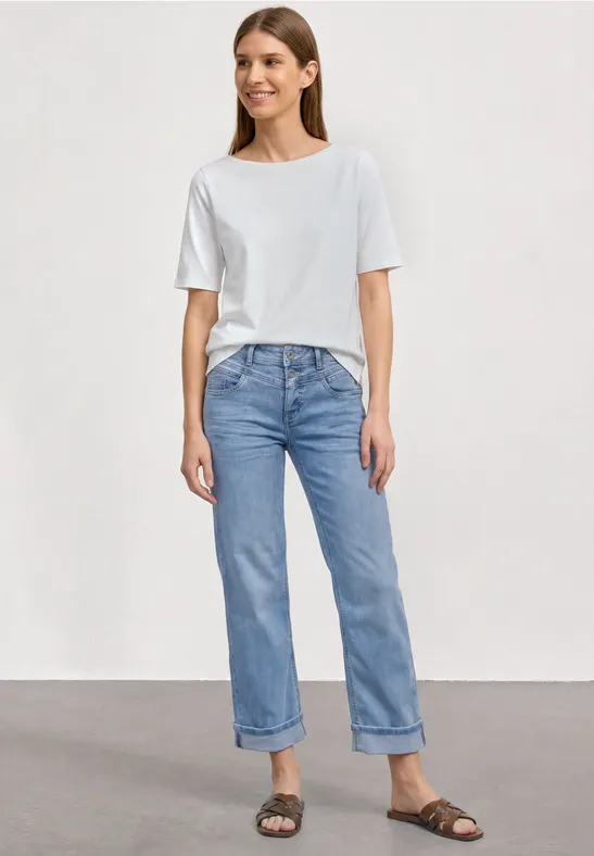 Vrouw draagt wit T-shirt, lichtblauwe opgerolde jeans en bruine sandalen, staand voor een neutrale achtergrond.