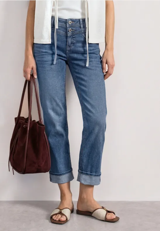 Vrouw draagt opgerolde blauwe spijkerbroek, wit topje, beige sandalen en bruine suède tas.