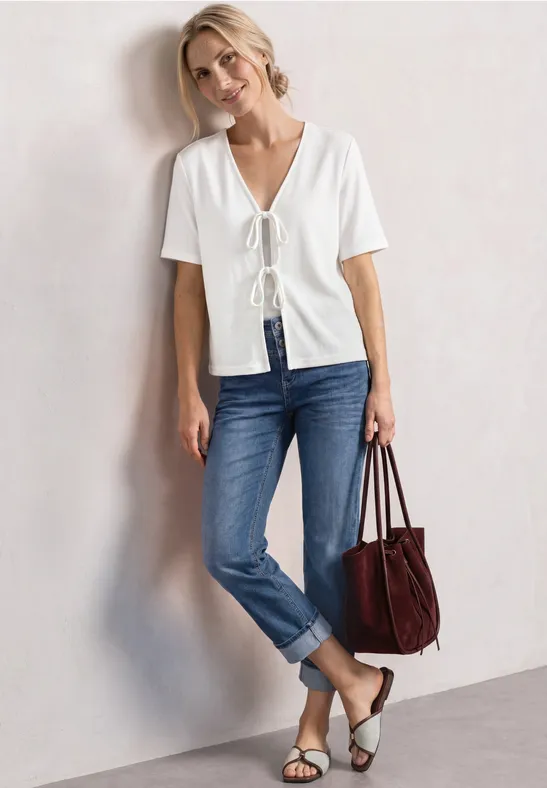 Vrouw leunt tegen muur in wit shirt met knoop, blauwe jeans, sandalen en rode tas.