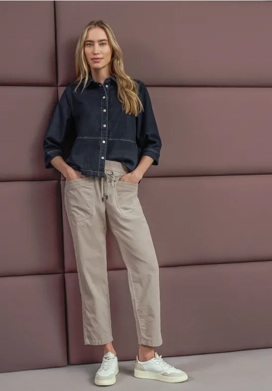 Een vrouw in een blauwe blouse, lichte broek en witte sneakers leunt ontspannen tegen een gestoffeerde wand.