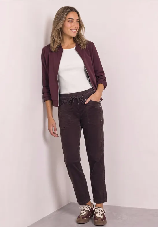 Een vrouw staat nonchalant in een hoek, draagt een wit T-shirt, een bordeauxblazer, donkere broek en sneakers.