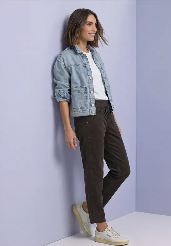 Een glimlachende vrouw in jeansjack, wit T-shirt en fluwelen broek leunt tegen een pastelkleurige muur.