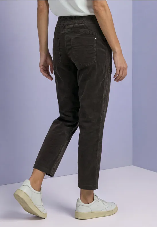 Achteraanzicht van persoon met zwarte corduroy broek en witte sneakers tegen paarse achtergrond.