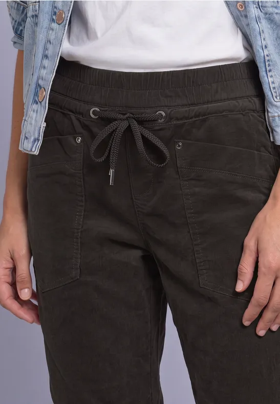 Close-up van een persoon met donkergroene corduroy broek met trekkoord en een denimvest over wit T-shirt.