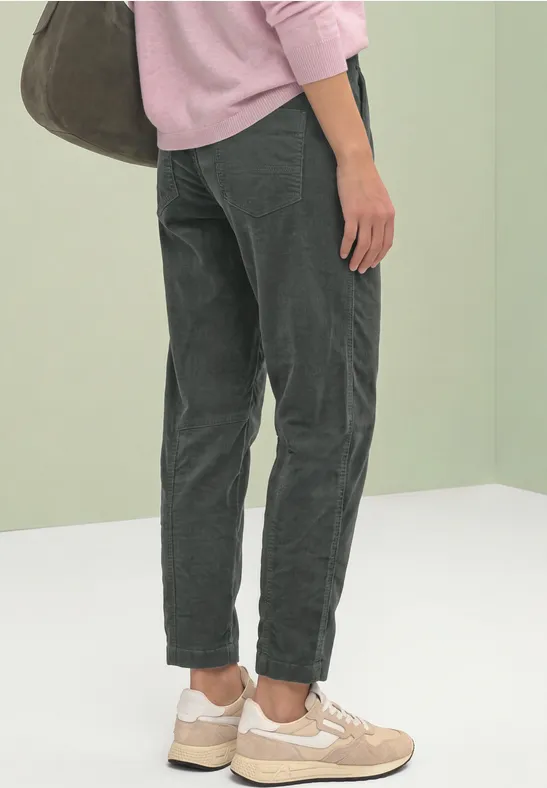 Vue de dos: personne en pantalon velours gris, baskets beiges, pull rose et grand sac.