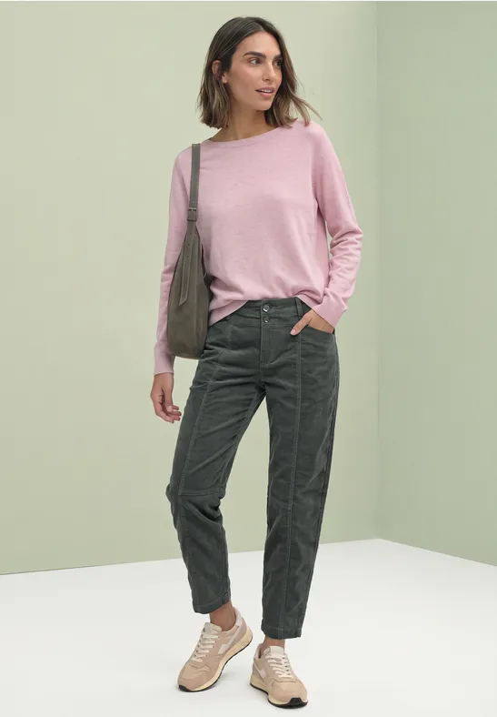 Femme en pull rose, pantalon cargo vert et baskets, portant un sac bandoulière devant un fond vert clair.