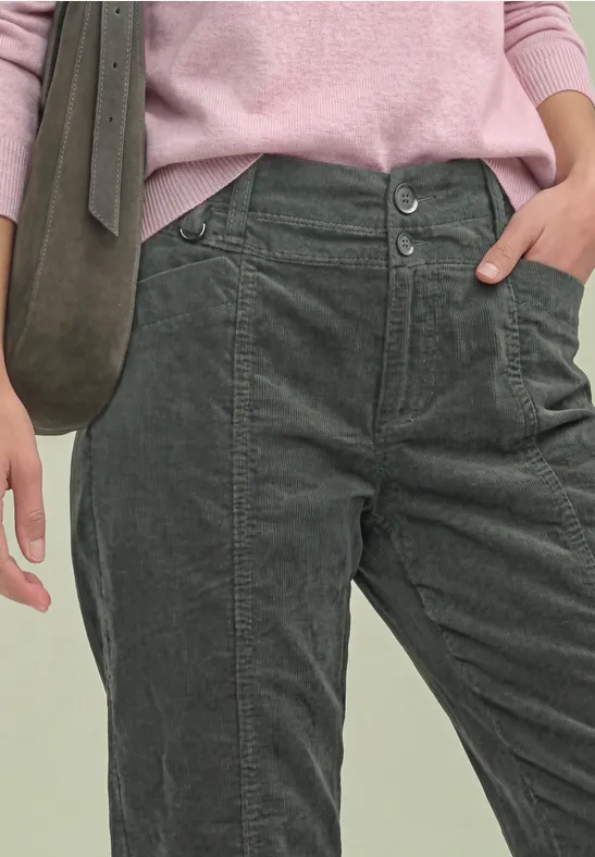Femme en pantalon velours côtelé vert-gris, pull rose et sac bandoulière vert (vue jusqu’à la taille).