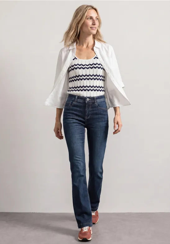 Femme portant une chemise blanche ouverte sur un top rayé, un jean bleu et des sandales.