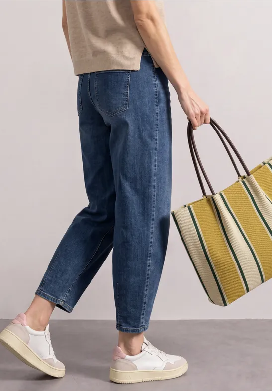 Vrouw in ruime jeans, witte sneakers en beige top, met een grote gestreepte tas.