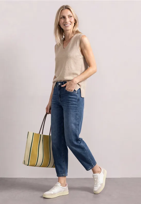 Een lachende vrouw in beige top, blauwe jeans en witte sneakers met een geel gestreepte tas.