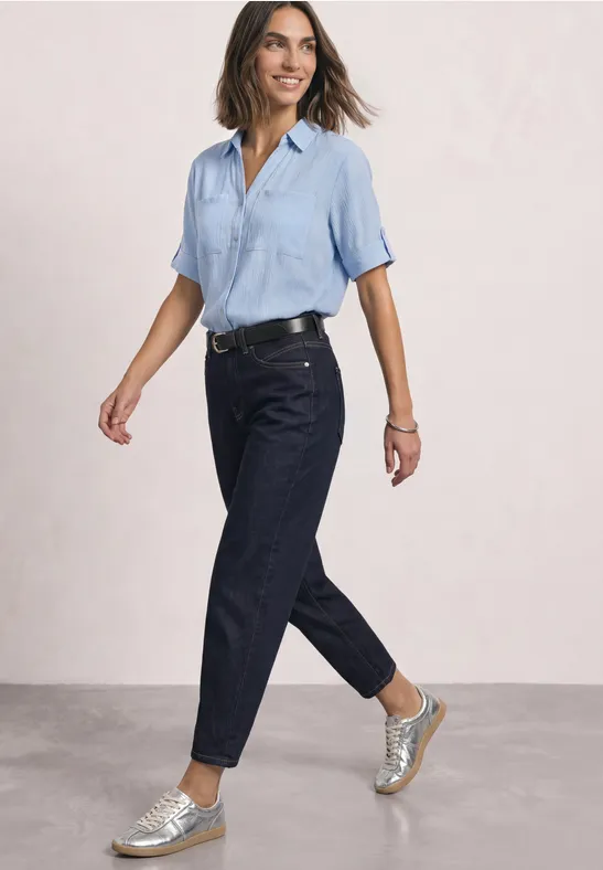 Een lachende vrouw in blauw hemd, donkere jeans en zilveren sneakers loopt voor een neutrale achtergrond.