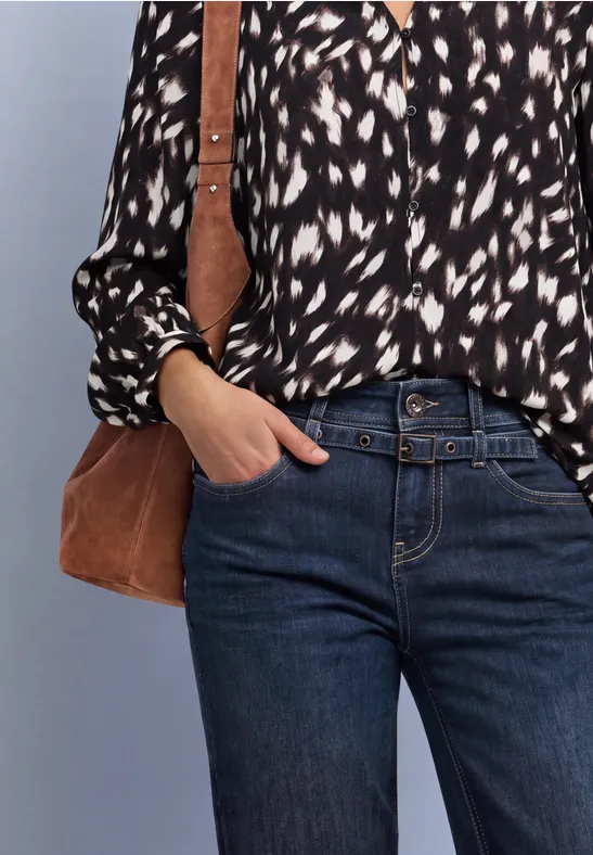 Vrouw met zwart-witblouse, blauwe spijkerbroek en bruine schoudertas, hand in de zak.