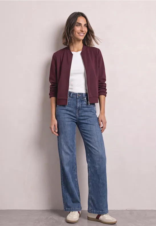 Femme en bomber bordeaux, t-shirt blanc, jean bleu taille haute et baskets blanches devant un mur clair