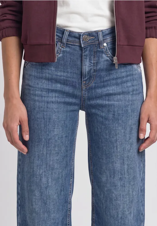 Femme portant un jean bleu et un sweat bordeaux, mains détendues le long du corps.