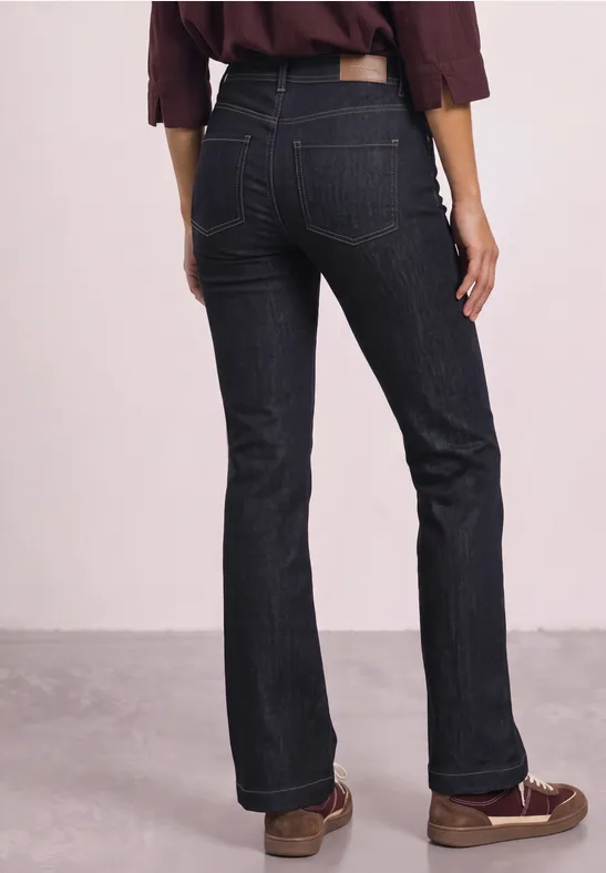 Femme de dos portant un jean taille haute sombre et des mocassins bruns sur fond neutre.