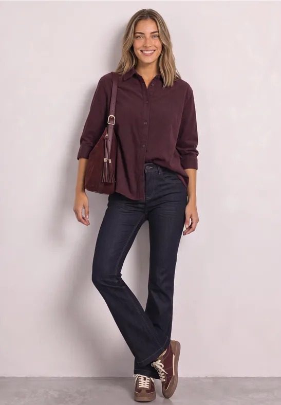 Une femme sur fond clair porte une chemise bordeaux, un jean noir, des baskets assorties et un sac à main.