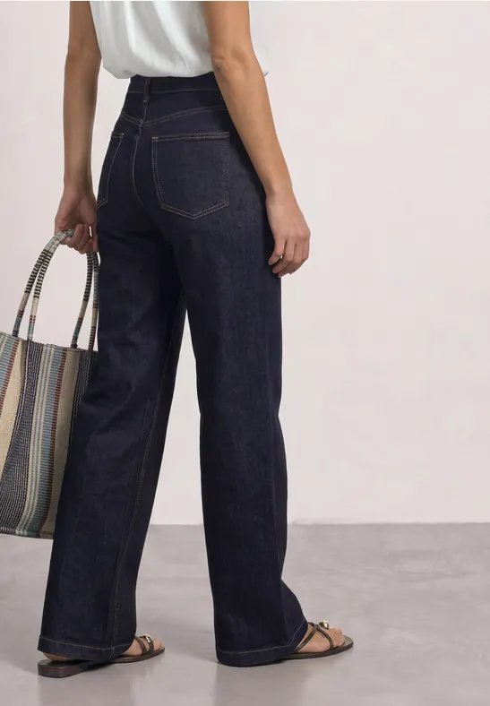 Vrouw van achteren in een hoge, wijde, donkerblauwe jeans, met sandalen en een gestreepte tas.