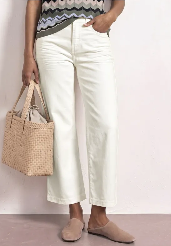 Een vrouw draagt crèmewitte, wijde cropped broek, platte instappers en houdt een gevlochten tas vast.