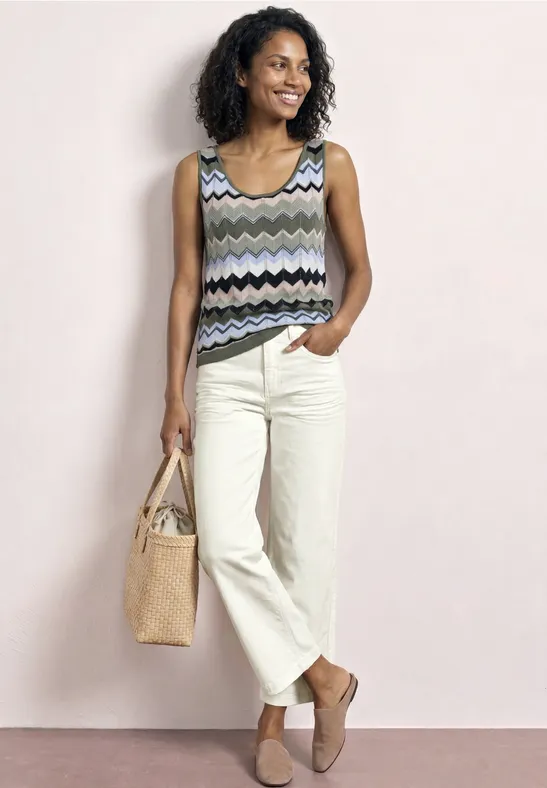 Een vrouw met krullend haar draagt een gestreept topje, lichte broek, rieten tas en platte schoenen.