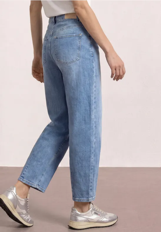 Vue latérale d'une personne portant un jean bleu clair ample et des baskets argentées sur fond uni.