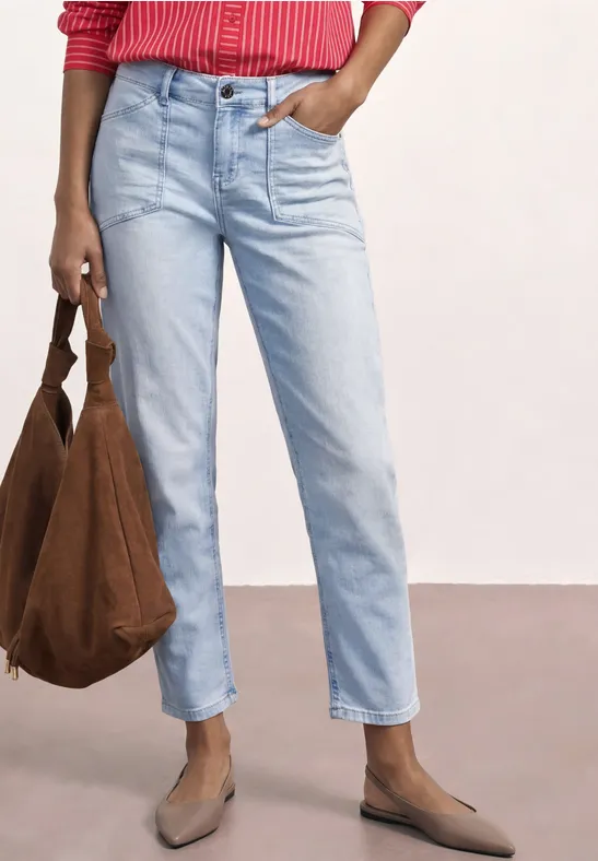 Eine Person in hellblauen Jeans, roter Bluse, hellbraunen Slippern und brauner Tasche vor hellem Hintergrund.