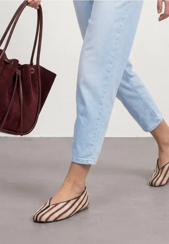 Frau in hellblauen Jeans trägt gestreifte Ballerinas und hält eine weinrote Wildledertasche.