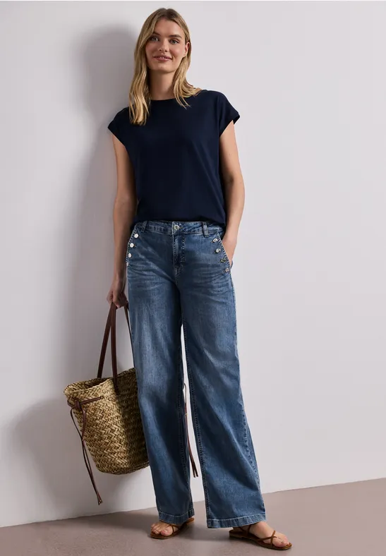 Frau in dunkelblauem T-Shirt, weiten Jeans, flachen Sandalen, mit geflochtenem Korb als Tasche.