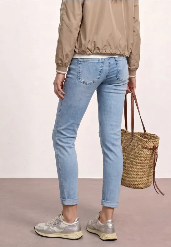 Vue de dos d'une personne en jeans bleu clair, baskets argentées, veste beige et sac en osier tressé.