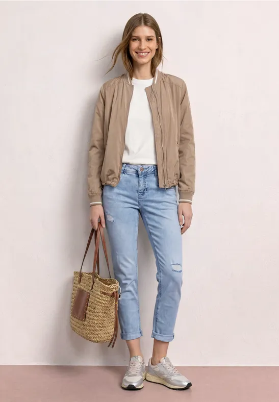 La femme porte une veste beige, un t-shirt blanc, un jean bleu et des baskets, tenant un sac en osier.