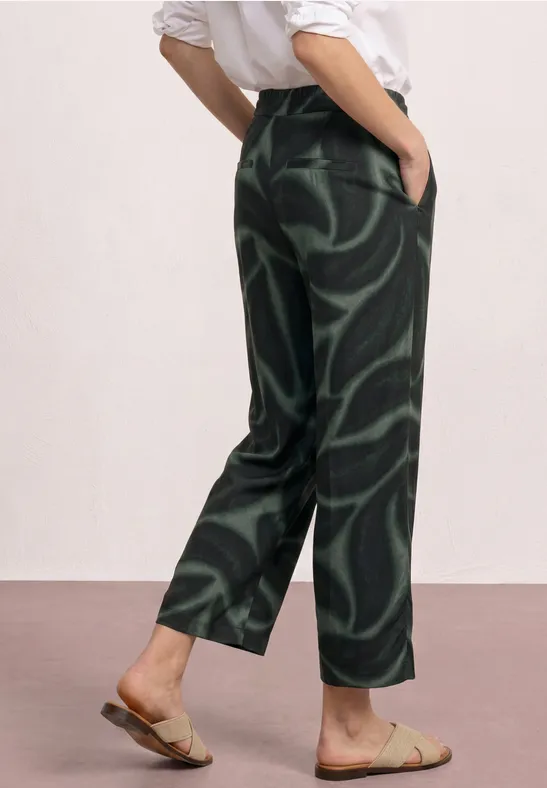 Persoon draagt een losse zwarte broek met groene wervelpatronen en platte sandalen.
