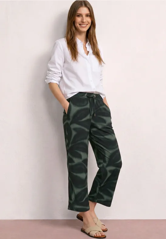 Een vrouw in een wit hemd en camouflerende broek staat voor een lichte muur.