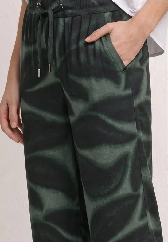 Close-up van een groene broek met zwarte golvende print, aantrekkoord en hand in de zak.
