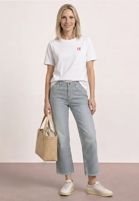 Une femme porte un t-shirt blanc, un jean clair, des baskets et tient un sac en paille devant un fond clair.